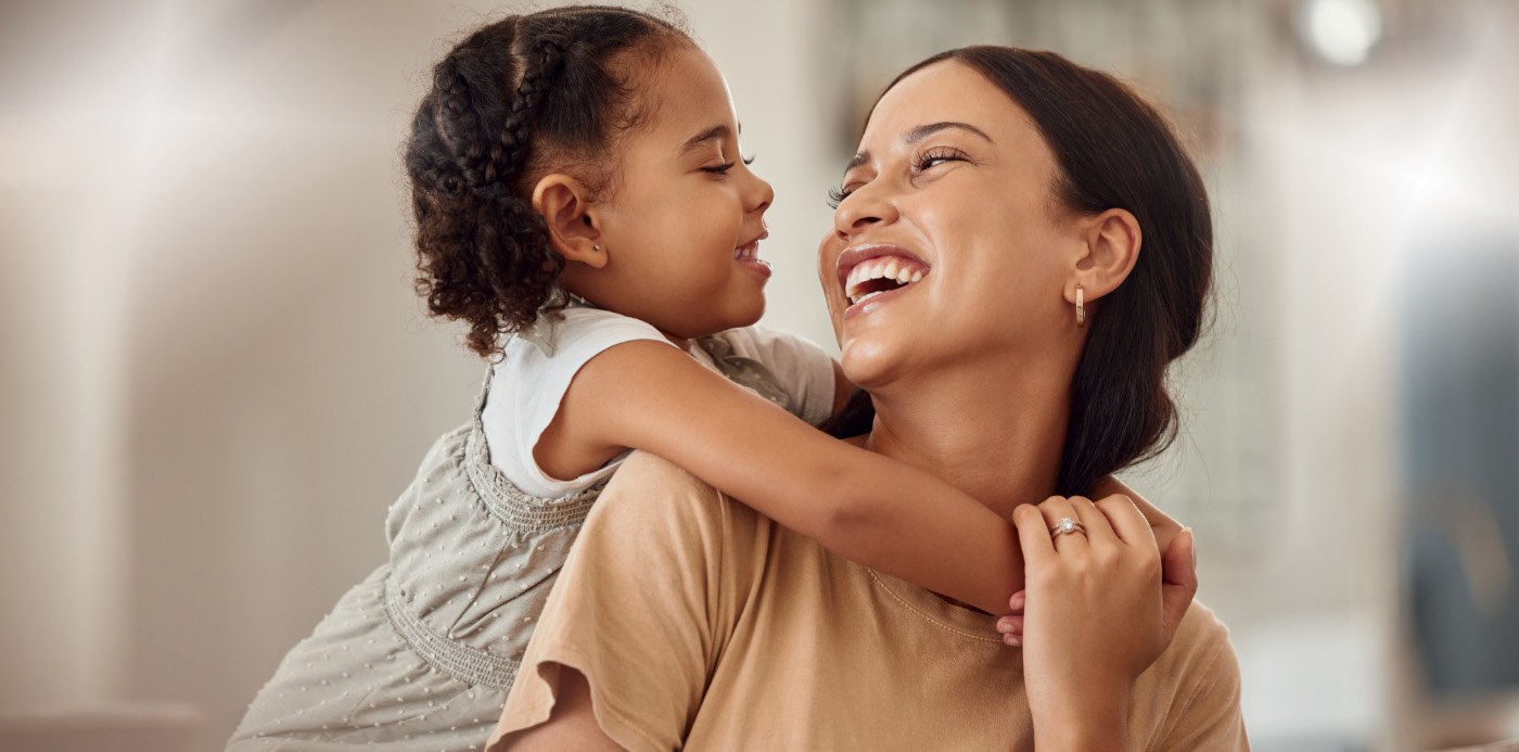 Smiling little girl hugging her mothers shoulders from behind